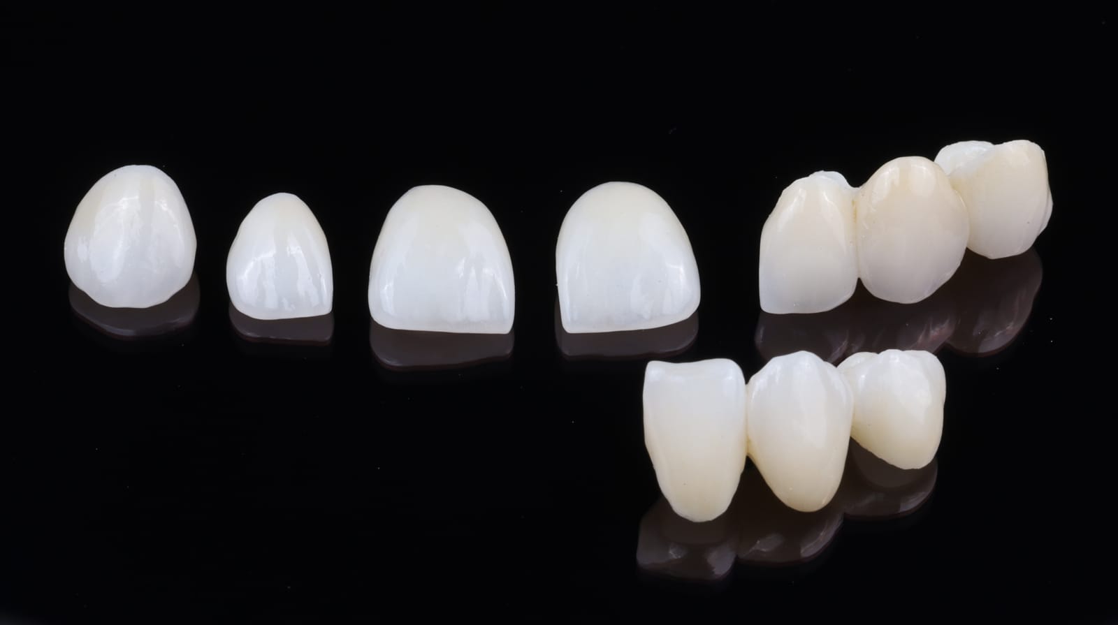 Ceramic crowns arranged on a black reflective surface, showing single anterior units alongside joined posterior bridge units.