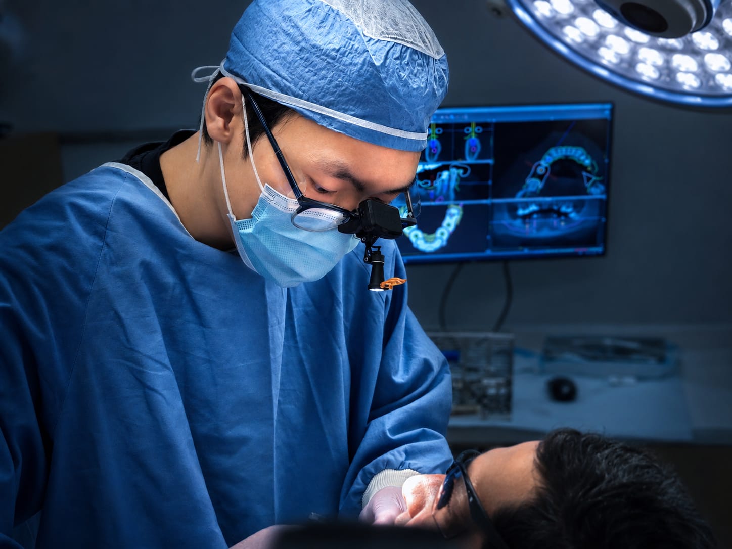 Dental implant surgery in progress at this practice. The operating clinician is in surgical cap, mask and magnification loupes, with the cone beam computed tomography planning scan on the monitor behind.